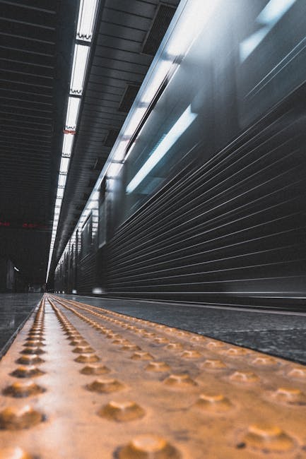 Inside a modern underground station corridor, viewed from a low angle close to the textured yellow tactile paving on the pavement. The corridor features metallic paneling along the walls and a dark ce