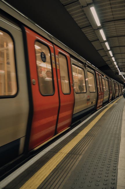 Inside a modern underground station corridor, viewed from a low angle close to the textured yellow tactile paving on the pavement. The corridor features metallic paneling along the walls and a dark ce